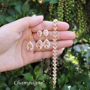 May include: A champagne-colored jewelry set featuring a necklace, earrings, and bracelet. The set includes teardrop-shaped pendants and earrings with a halo of clear stones. The bracelet is adorned with similar stones. The word "Champagne" is written in the image.