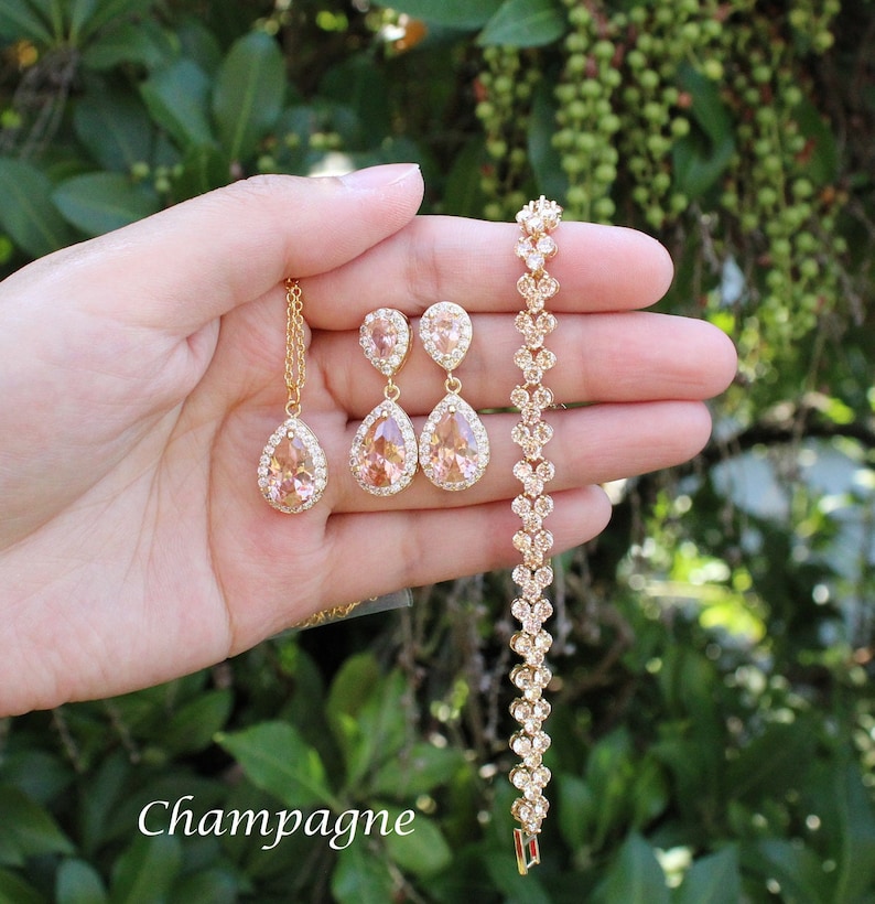 May include: A champagne-colored jewelry set featuring a necklace, earrings, and bracelet. The set includes teardrop-shaped pendants and earrings with a halo of clear stones. The bracelet is adorned with similar stones. The word "Champagne" is written in the image.