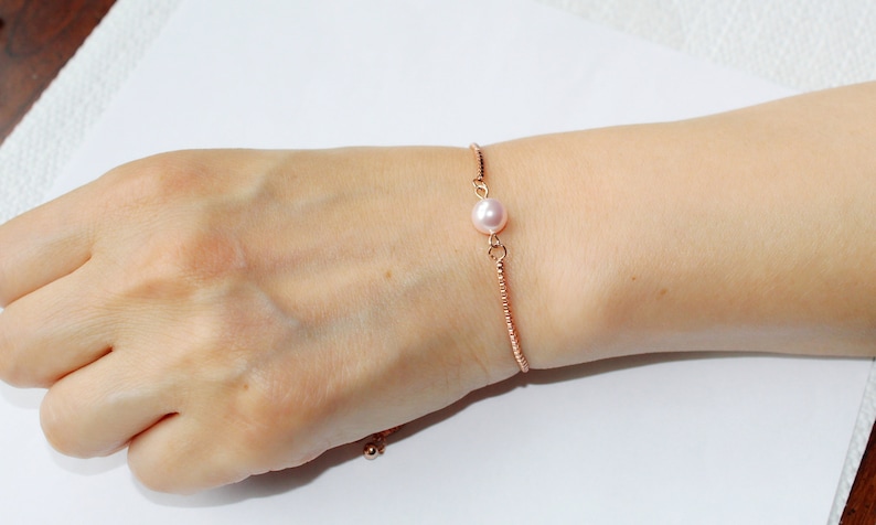 May include: A delicate rose gold bracelet with a single pink pearl charm. The bracelet is adjustable and features a sliding knot closure.