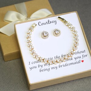 Puede incluir: Una caja de regalo dorada con una tarjeta blanca en su interior. La tarjeta tiene el texto "Courtney" y un mensaje que dice "No podr&iacute;a casarme sin ti a mi lado. Gracias por ser mi dama de honor!" La caja contiene un brazalete dorado y un par de pendientes con piedras transparentes.