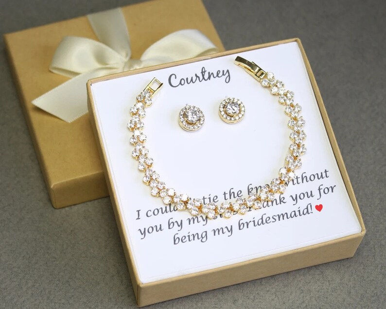 Puede incluir: Una caja de regalo dorada con una tarjeta blanca en su interior. La tarjeta tiene el texto "Courtney" y un mensaje que dice "No podr&iacute;a casarme sin ti a mi lado. Gracias por ser mi dama de honor!" La caja contiene un brazalete dorado y un par de pendientes con piedras transparentes.