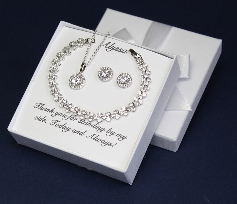 Puede incluir: Una caja de regalo blanca con un conjunto de collar, pulsera y pendientes de plata. La caja tiene el texto "Gracias por estar a mi lado. Hoy y siempre!" y el nombre "Alyssa" escrito en ella.