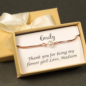 May include: A small gift box with a white satin ribbon and a personalized message inside. The message reads "Emily, Thank you for being my flower girl! Love, Madison". A delicate rose gold bracelet with a single pearl charm is inside the box.