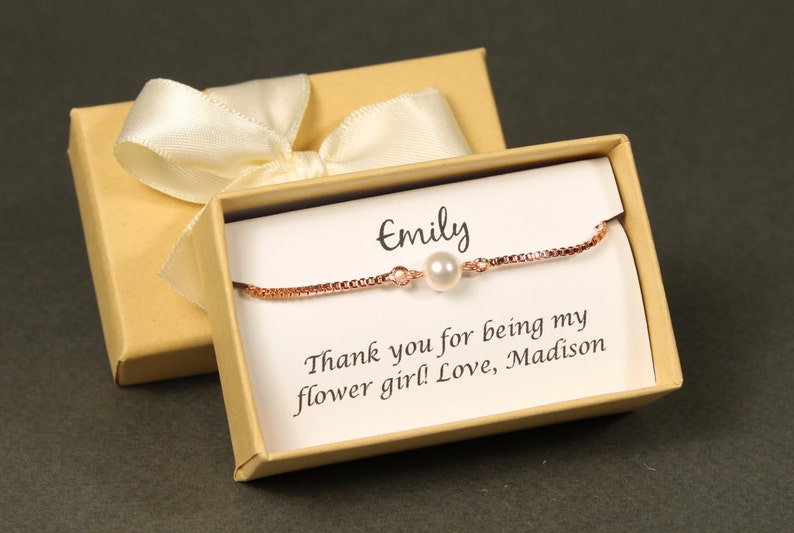 May include: A small gift box with a white satin ribbon and a personalized message inside. The message reads "Emily, Thank you for being my flower girl! Love, Madison". A delicate rose gold bracelet with a single pearl charm is inside the box.