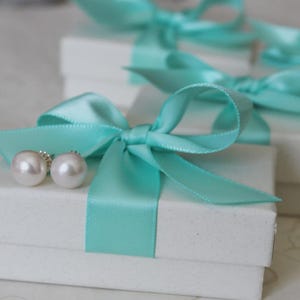May include: Two white round pearl stud earrings resting on a small white rectangular cardboard gift box. The box is tied with a bright turquoise satin ribbon bow. Additional identical gift boxes are visible in the blurred background.