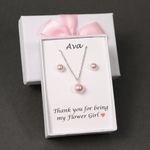 May include: A white gift box with a pink ribbon and a white card inside. The card has the name "Ava" printed on it and says "Thank you for being my Flower Girl". The box contains a pink pearl necklace and earrings.