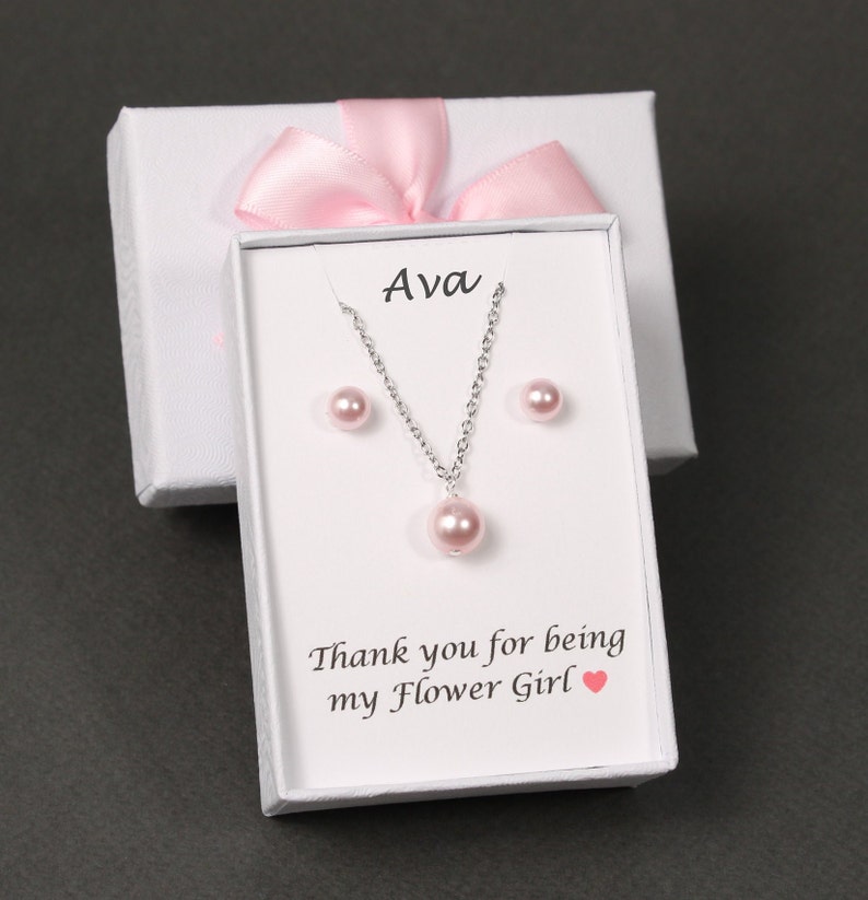 May include: A white gift box with a pink ribbon and a white card inside. The card has the name "Ava" printed on it and says "Thank you for being my Flower Girl". The box contains a pink pearl necklace and earrings.