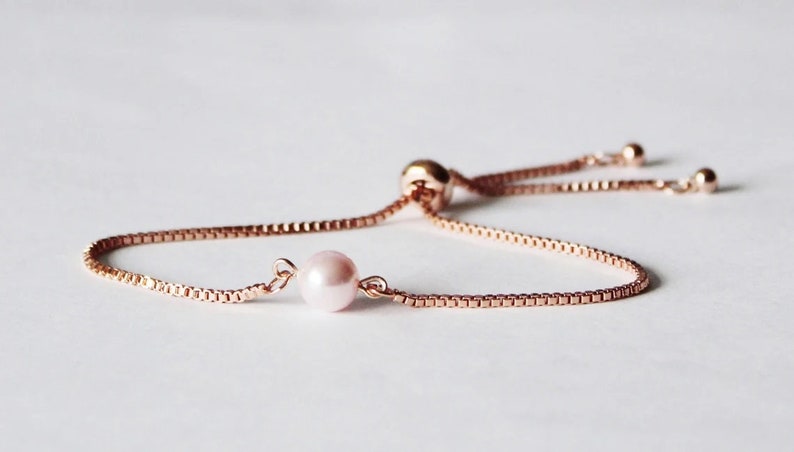 May include: A delicate rose gold chain bracelet with a single pink pearl charm. The bracelet has an adjustable sliding knot closure.