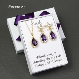 May include: A white gift box with a purple ribbon contains a gold necklace and earrings set with a teardrop-shaped purple gemstone and clear crystals. The box has a card inside that reads "Sarah Thank you for standing by my side. Today and Always!"