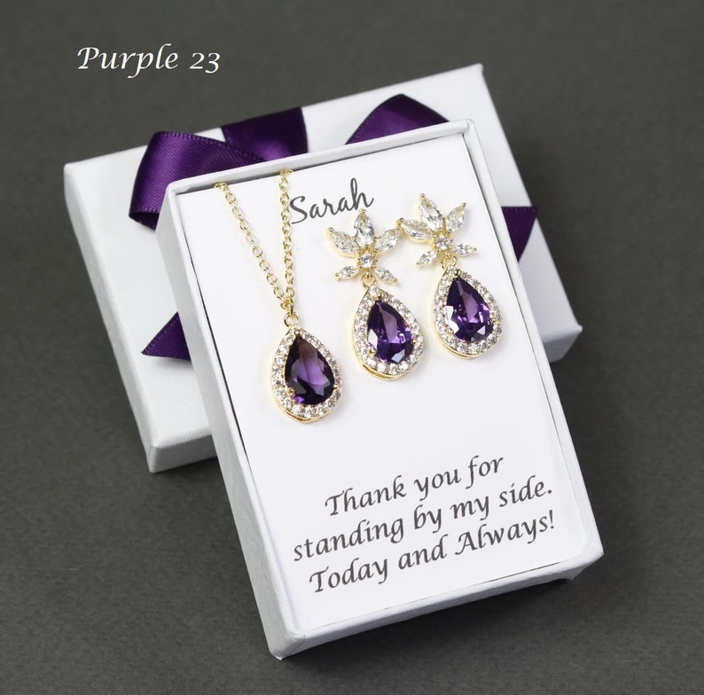 May include: A white gift box with a purple ribbon contains a gold necklace and earrings set with a teardrop-shaped purple gemstone and clear crystals. The box has a card inside that reads "Sarah Thank you for standing by my side. Today and Always!"