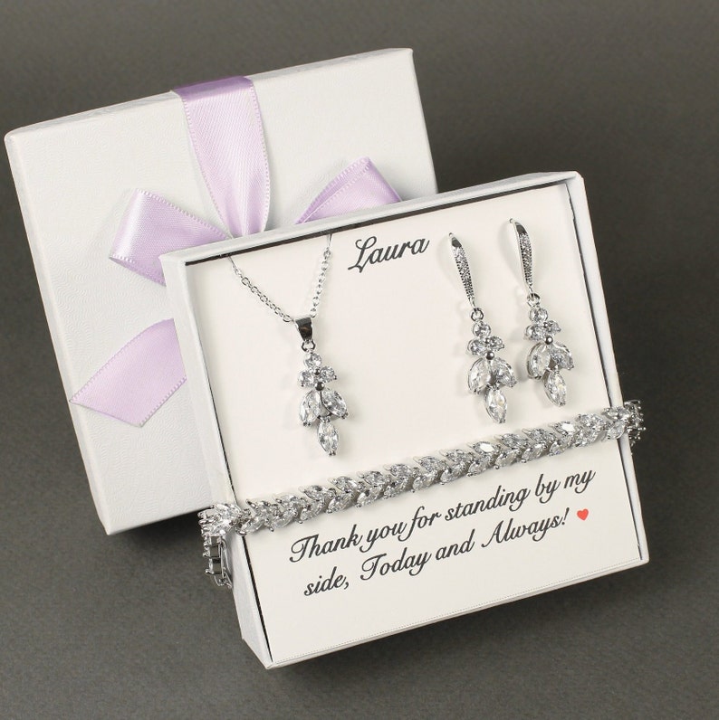 May include: White gift box with a lavender ribbon, containing a silver necklace, earrings, and bracelet with clear crystal stones. The box has a personalized message that reads "Thank you for standing by my side, Today and Always!"
