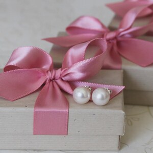 May include: Two white pearl stud earrings resting on a rectangular beige cardboard gift box. The box is tied with a wide, shiny pink satin ribbon tied into a bow. A second identical gift box is visible in the background, also featuring a pink bow.
