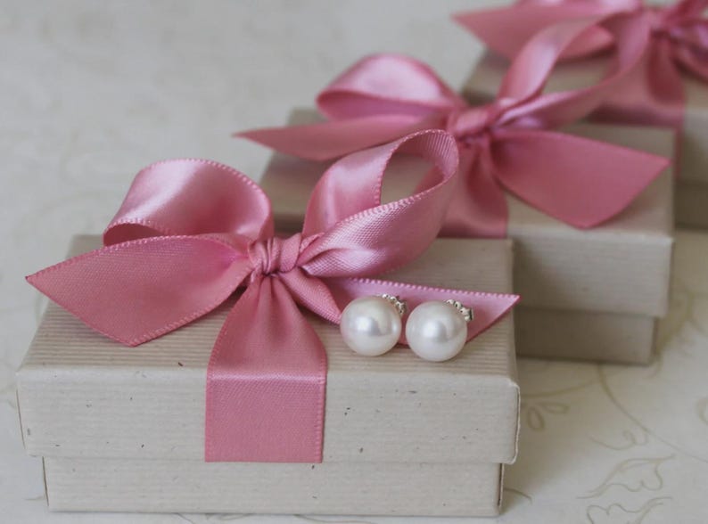 May include: Two white pearl stud earrings resting on a rectangular beige cardboard gift box. The box is tied with a wide, shiny pink satin ribbon tied into a bow. A second identical gift box is visible in the background, also featuring a pink bow.