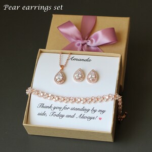 Puede incluir: Una caja de regalo con una cinta rosa contiene un collar, pendientes y una pulsera de oro rosa con piedras transparentes en forma de pera. La caja tiene un mensaje personalizado que dice "Amanda Gracias por estar a mi lado, hoy y siempre!"