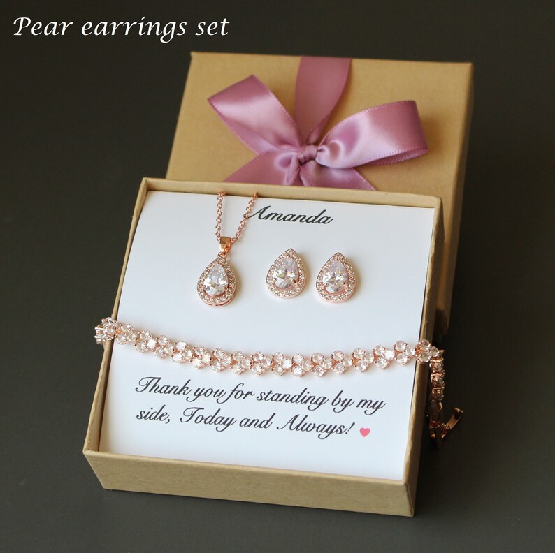 Puede incluir: Una caja de regalo con una cinta rosa contiene un collar, pendientes y una pulsera de oro rosa con piedras transparentes en forma de pera. La caja tiene un mensaje personalizado que dice "Amanda Gracias por estar a mi lado, hoy y siempre!"