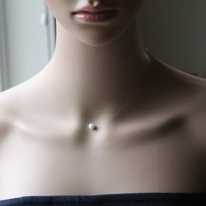 May include: A delicate, single-pearl necklace on a clear, nearly invisible string. The pearl is white and round, hanging on the center of the chest.