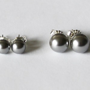 May include: Two pairs of gray pearl stud earrings. The earrings have a round shape and are set on silver posts. The pearls have a smooth, glossy finish. The earrings are displayed on a white surface.