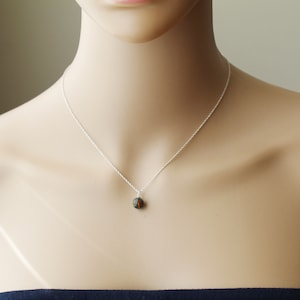 以下が含まれることがあります： 小さな丸いペンダントが付いたシルバーのネックレス。ペンダントは濃い緑色と赤色の混合色です。ネックレスはマネキンに展示されています。