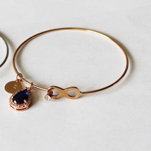 May include: A rose gold bangle bracelet with a charm featuring a blue teardrop-shaped stone surrounded by smaller stones and an infinity symbol.