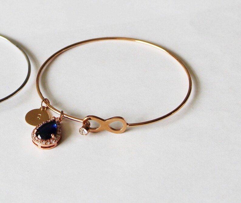 May include: A rose gold bangle bracelet with a charm featuring a blue teardrop-shaped stone surrounded by smaller stones and an infinity symbol.