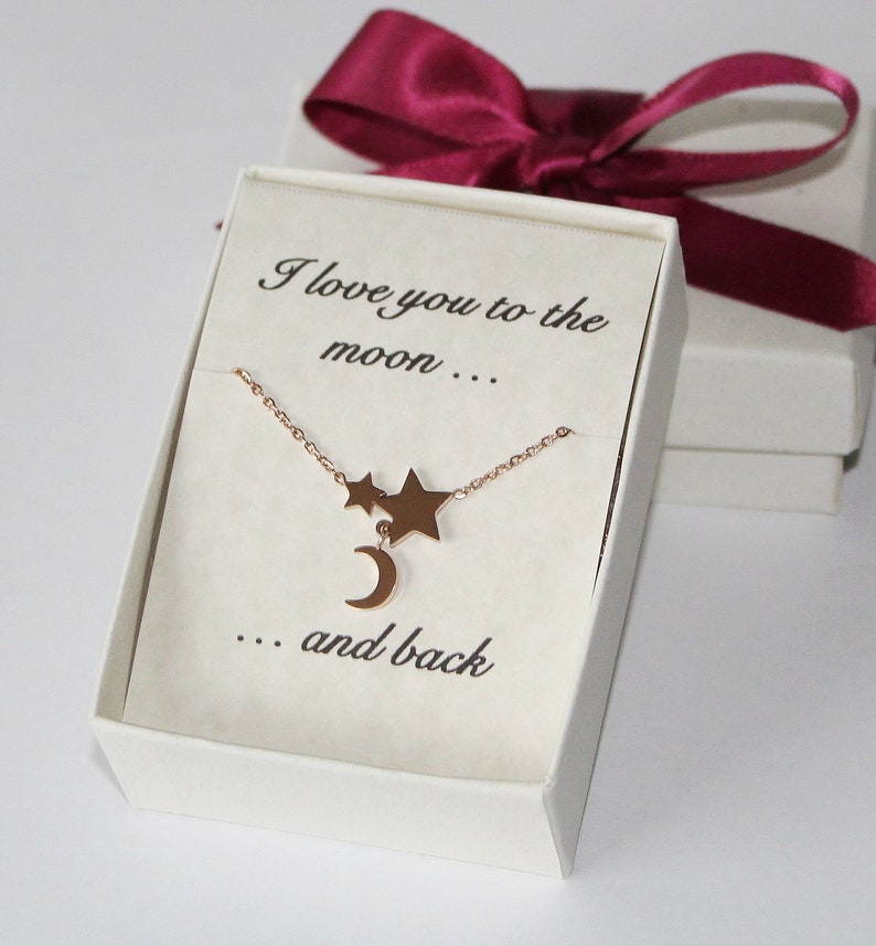 May include: A white gift box with a red ribbon and a gold necklace with a crescent moon and two stars charm. The box has the text "I love you to the moon... ...and back" printed on it.