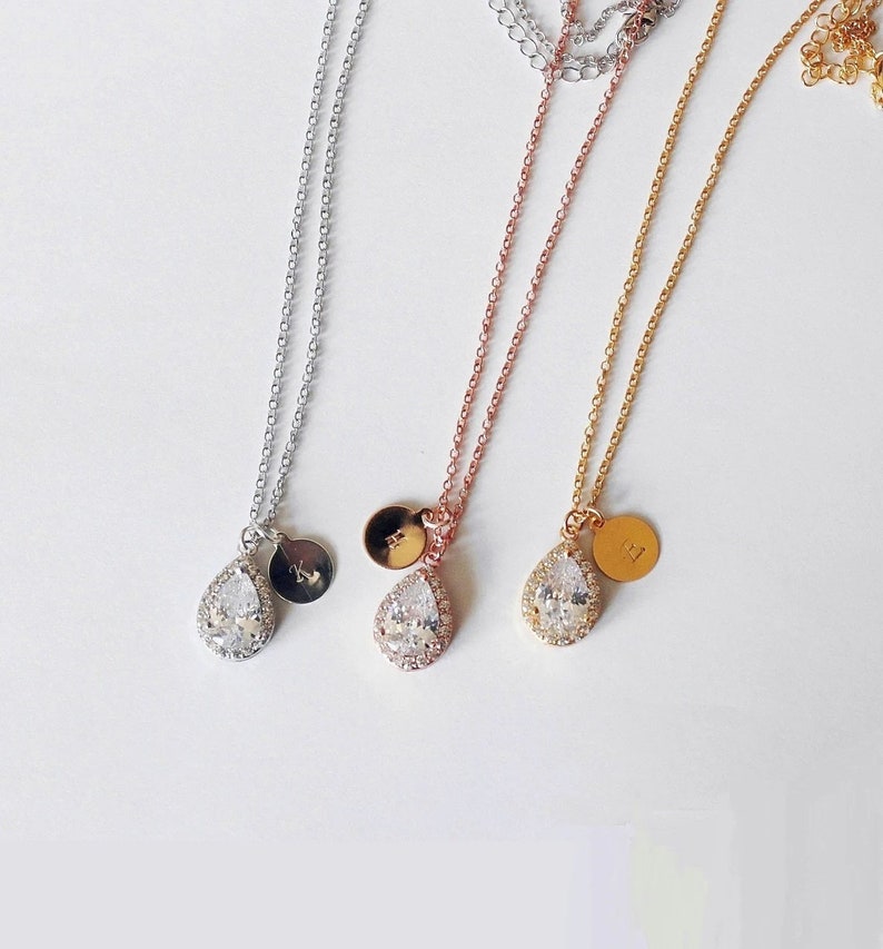 May include: Three delicate necklaces in silver, rose gold, and gold. Each features a teardrop-shaped pendant with a halo of clear stones and a small, round charm with an initial. The chains are fine and the pendants are centered.