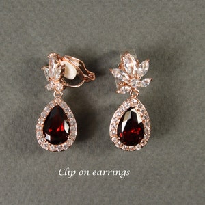 May include: A pair of clip-on earrings with a teardrop-shaped red stone surrounded by clear rhinestones. The earrings have a floral design at the top.