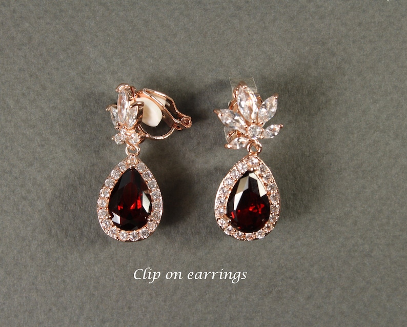 May include: A pair of clip-on earrings with a teardrop-shaped red stone surrounded by clear rhinestones. The earrings have a floral design at the top.