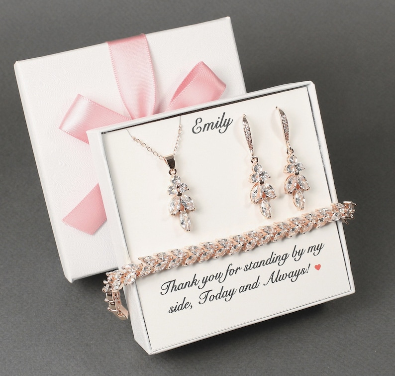 May include: A white gift box with a pink ribbon and a smaller white box inside. The smaller box contains a rose gold necklace, earrings, and bracelet with clear crystal accents. The smaller box also has the name "Emily" and the text "Thank you for standing by my side, Today and Always!" printed on it.