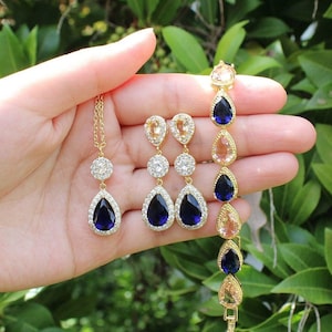 Puede incluir: Un conjunto de joyas doradas con piedras azules y melocotón en forma de lágrima. El conjunto incluye un collar, pendientes y una pulsera.