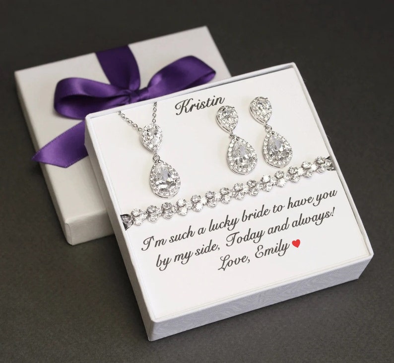 May include: A white gift box with a purple ribbon and a message inside. The message reads "I'm such a lucky bride to have you by my side, Today and always! Love, Emily". The box contains a silver necklace, earrings, and bracelet with teardrop-shaped crystals.