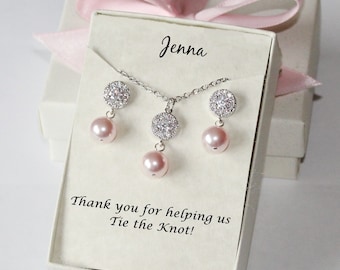 Puede incluir: Una caja de regalo blanca con una cinta rosa y una tarjeta blanca en su interior. La tarjeta tiene el nombre "Jenna" escrito en ella y el texto "¡Gracias por ayudarnos a atar el nudo!" La caja contiene un collar y pendientes de plata con detalles de perlas rosas y adornos de cristal transparente.