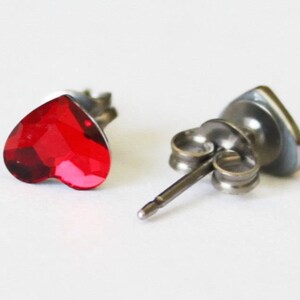 May include: A pair of silver heart-shaped stud earrings with red crystal accents.