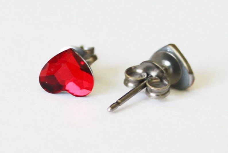 May include: A pair of silver heart-shaped stud earrings with red crystal accents.