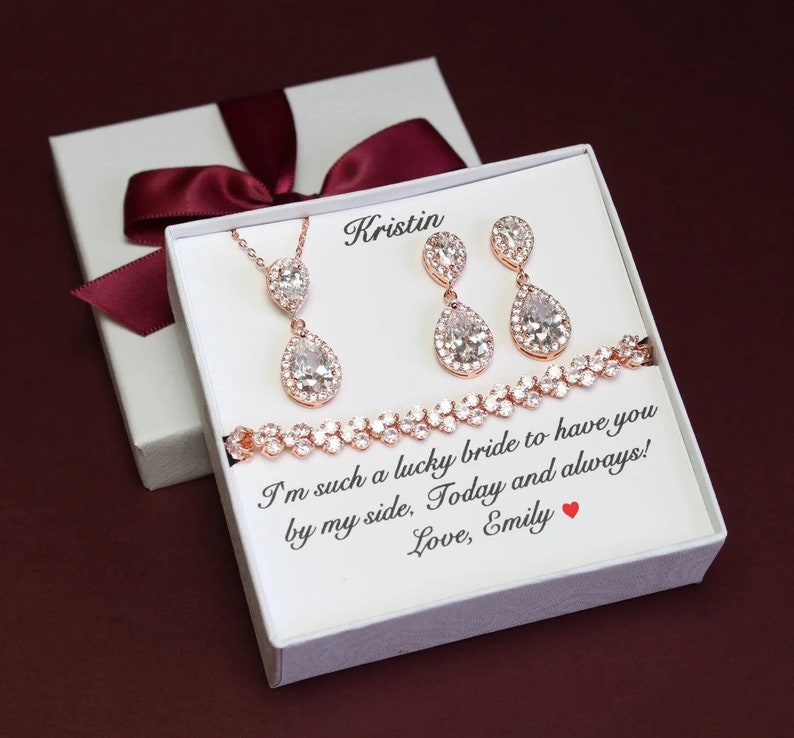 May include: A white gift box with a burgundy ribbon contains a rose gold necklace, earrings, and bracelet with teardrop-shaped crystals. The box has a personalized message that reads "Kristin I'm such a lucky bride to have you by my side, Today and always! Love, Emily"
