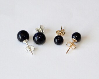 May include: Four stud earrings featuring spherical dark blue sandstone beads with shimmering flecks. Two earrings have silver-toned metal posts and butterfly backs, while the other two earrings have gold-toned metal posts and butterfly backs.