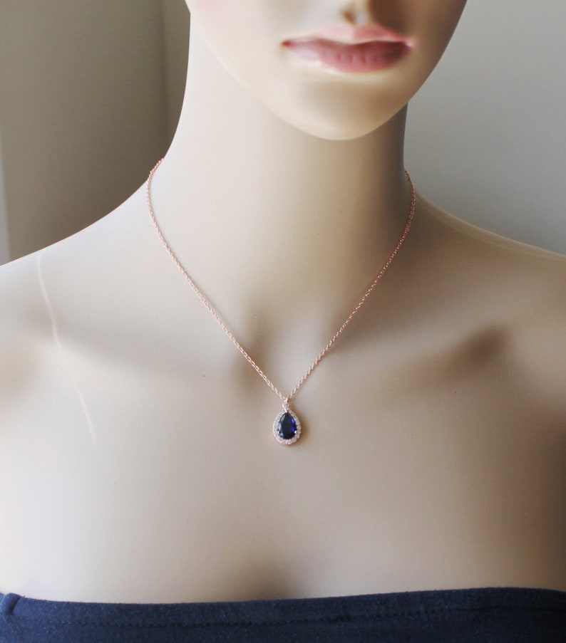 May include: A rose gold necklace with a teardrop-shaped pendant featuring a dark blue sapphire surrounded by a halo of smaller clear stones.
