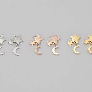 May include: Three pairs of earrings with a crescent moon charm dangling from a cluster of three stars. The earrings are shown in silver, rose gold, and gold.