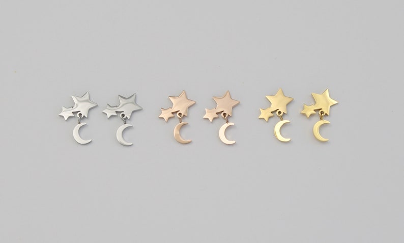 May include: Three pairs of earrings with a crescent moon charm dangling from a cluster of three stars. The earrings are shown in silver, rose gold, and gold.