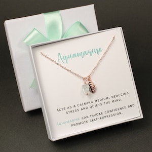 May include: A delicate rose gold necklace with a small aquamarine stone and a leaf charm. The necklace comes in a white gift box with a light green ribbon and a card that reads "Aquamarine ACTS AS A CALMING MEDIUM, REDUCING STRESS AND QUIETS THE MIND. AQUAMARINE CAN INVOKE CONFIDENCE AND PROMOTE SELF-EXPRESSION."