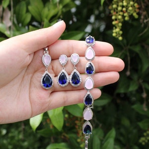 May include: A set of teardrop-shaped earrings and a bracelet with alternating pink and blue stones. The earrings have a silver setting and the stones are a light pink and a dark blue. The bracelet has a silver chain and the stones are a light pink and a dark blue.