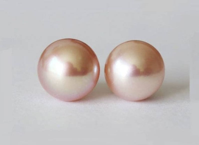May include: A pair of round, smooth, light pink pearls with a lustrous, reflective surface. Each pearl exhibits a soft, iridescent sheen and a uniform spherical shape, showcasing a delicate pastel color palette and a polished, high-gloss finish.