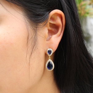 May include: Gold-toned drop earrings featuring a teardrop-shaped, deep blue gemstone. The gemstones are surrounded by a halo of small, clear crystals. The earrings are dangling from a smaller, similar design.
