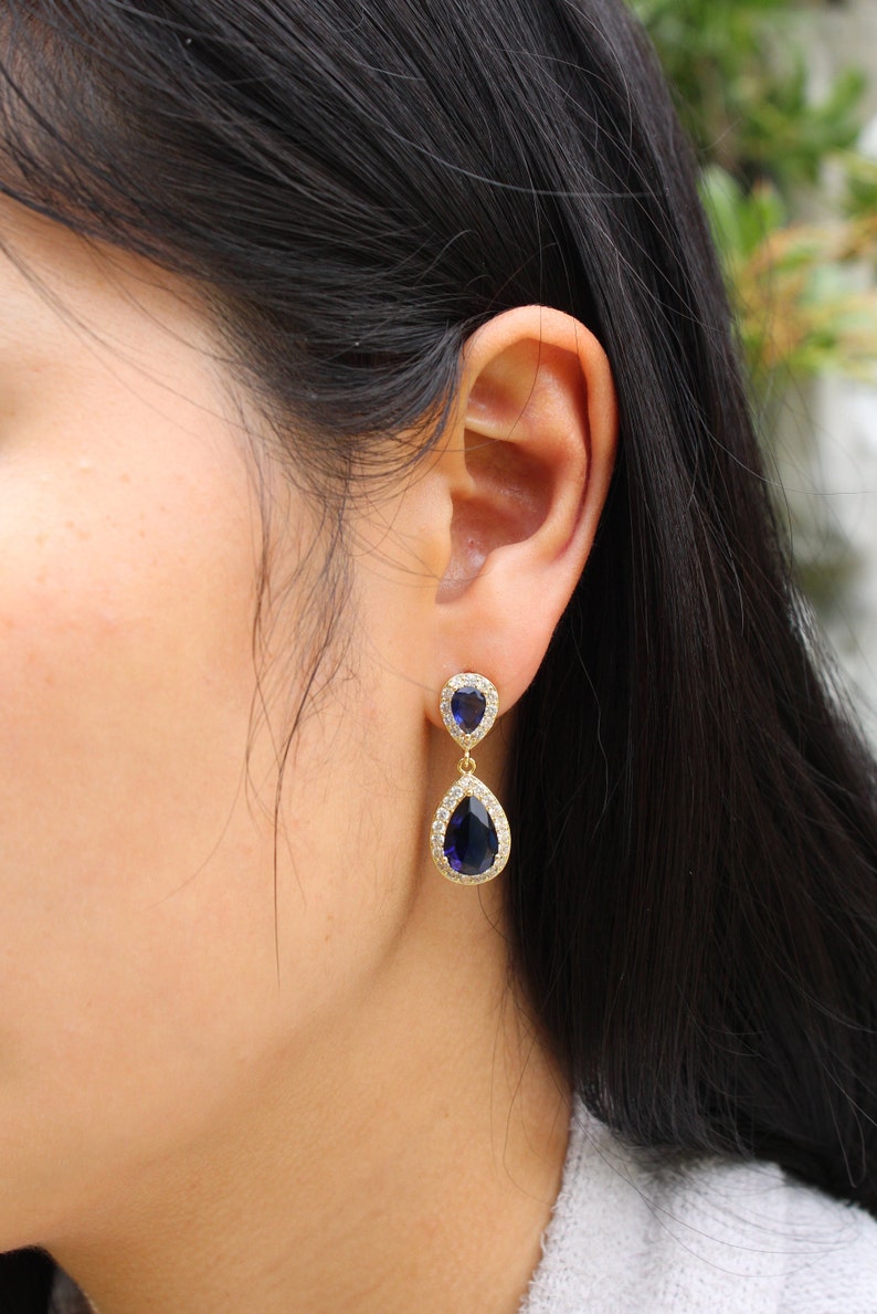 May include: Gold-toned drop earrings featuring a teardrop-shaped, deep blue gemstone. The gemstones are surrounded by a halo of small, clear crystals. The earrings are dangling from a smaller, similar design.