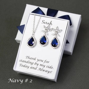 May include: A white gift box with a blue ribbon, containing a silver necklace and earrings with teardrop-shaped blue stones. The box has a personalized message that reads "Sarah Thank you for standing by my side. Today and Always!" and "Navy # 2".