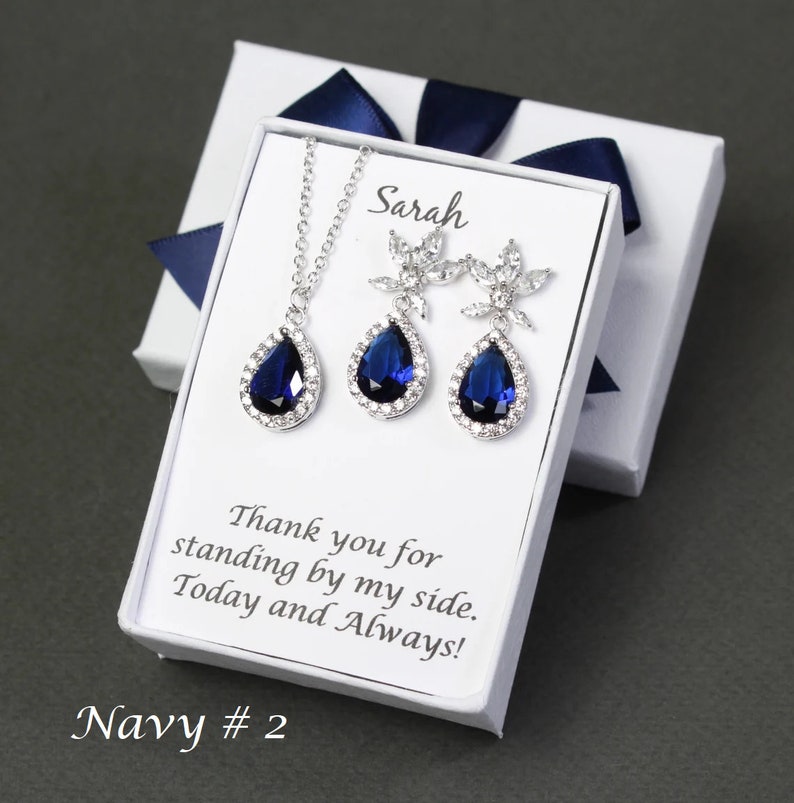 May include: A white gift box with a blue ribbon, containing a silver necklace and earrings with teardrop-shaped blue stones. The box has a personalized message that reads "Sarah Thank you for standing by my side. Today and Always!" and "Navy # 2".