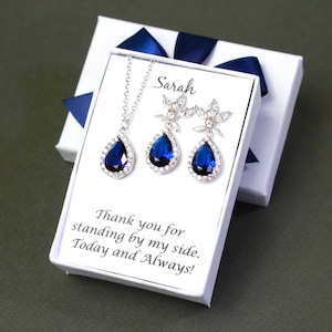 May include: A white gift box with a blue ribbon contains a blue teardrop necklace and earrings set. The box has a personalised message that reads "Sarah" and "Thank you for standing by my side. Today and Always!"