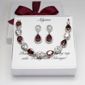 May include: A white box with a red bow on top. Inside the box is a silver bracelet and earrings set with red and clear stones. The box has the text "Alyssa" and "Than by my side. Today and Always!" printed on it.