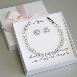 May include: A white gift box with a pink ribbon and a silver bracelet and earrings set with clear stones. The box has the name "Alyssa" written on it and the text "Thank you for standing by my side. Today and Always!" inside.