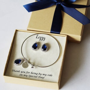 May include: A gift box with a blue ribbon and a personalized message for "Lizzy". Inside the box is a silver bangle bracelet with a teardrop-shaped blue stone charm and a pair of matching earrings.